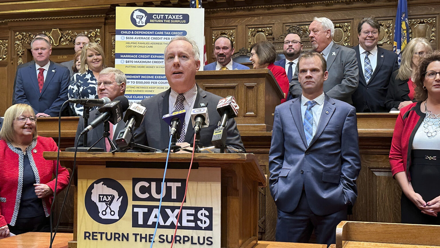 Robin Vos speaks into multiple microphones with the flags of different media organizations mounted to a wood podium with a sign on its front reading Cut Taxe$ and Return the Surplus that is part at the front of a legislative dais, with Devin LeMahieu standing to one side and other people standing on either side and on a higher tier, with a sign with the same title and multiple bullet points and a Wisconsin flag in the background of a room with carved wood wall panels.
