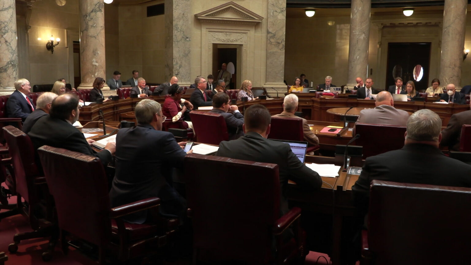 State senators sit at wood desks arranged in a semi-circle in a room with marble masonry and pillars.