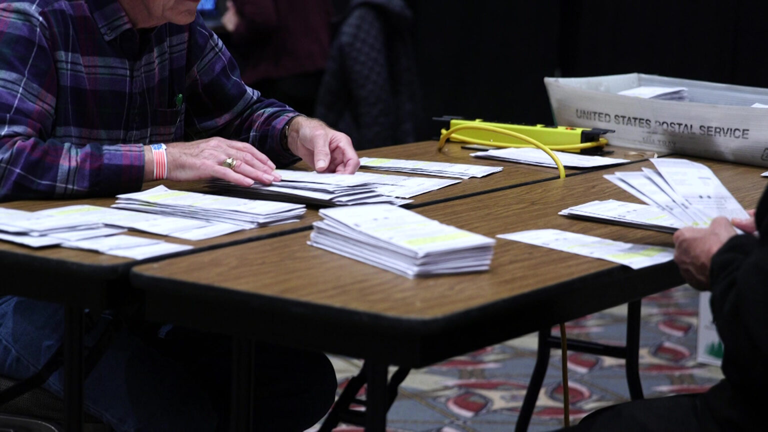 Two poll workers sitting on opposite sides of two folding tables with laminate wood surfaces placed side-by-side handle absentee ballots, with multiple stacks of ballots atop the table alongside a empty plastic U.S. mail trays.