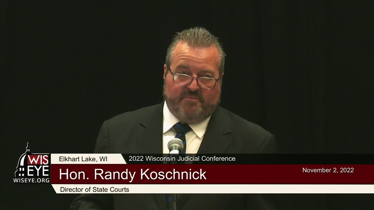 A video still image shows Randy Koschnick speaking into a microphone in front of a stage curtain, with a video graphic at bottom including the text 2022 Wisconsin Judicial Conference and Director of State Courts.