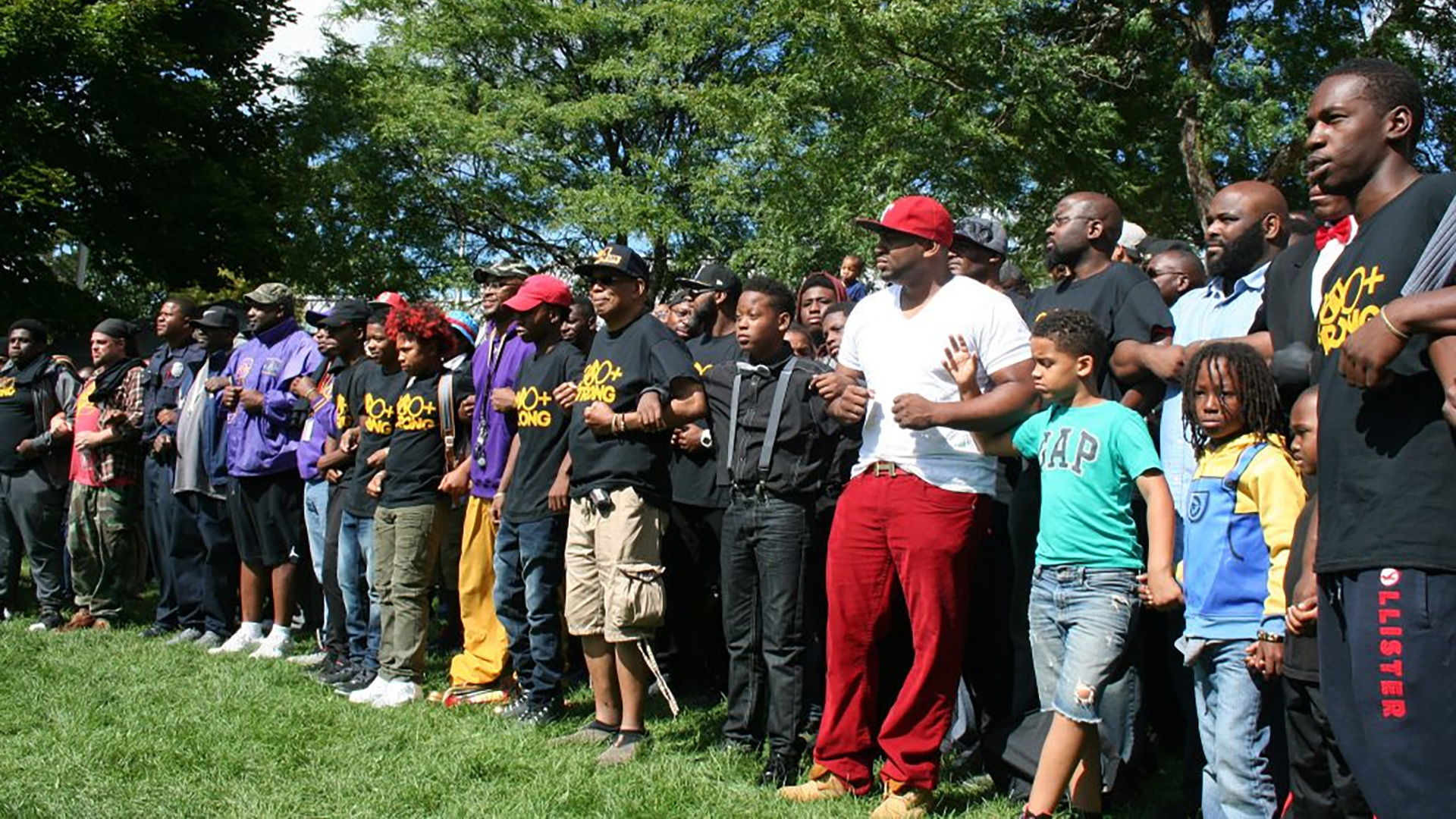 Dozens of men and boys stand in a line with their arms linked, with trees in the background.