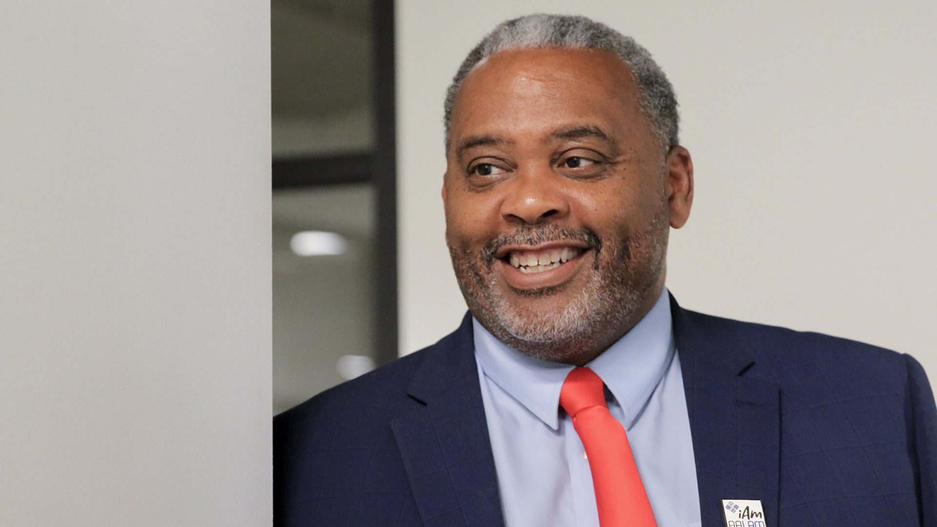 Walter Lanier smiles while standing near the exterior corner of walls in a room with blank walls and a window reflecting fluorescent lights.