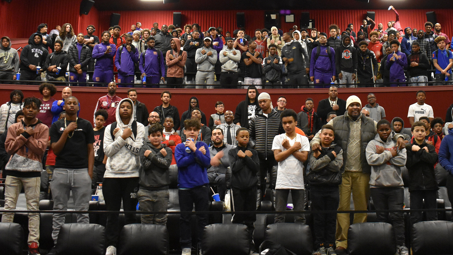 Dozens of male students stand in a movie theater with tiered seating in two levels while holding their arms crossed over their chests, with a row of empty seats at the bottom.