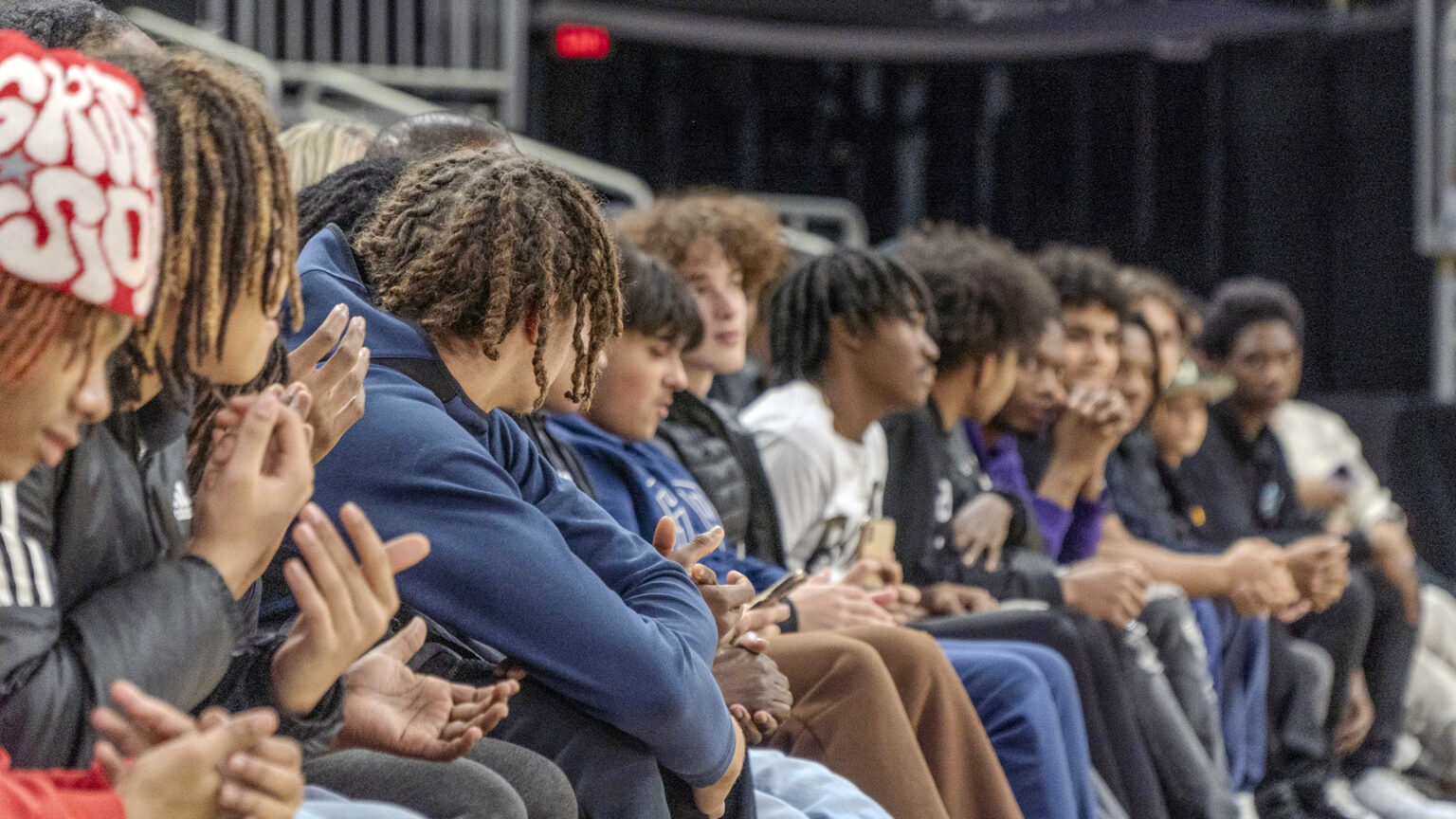 More than a dozen male students listen while seated next to each other on a bench.