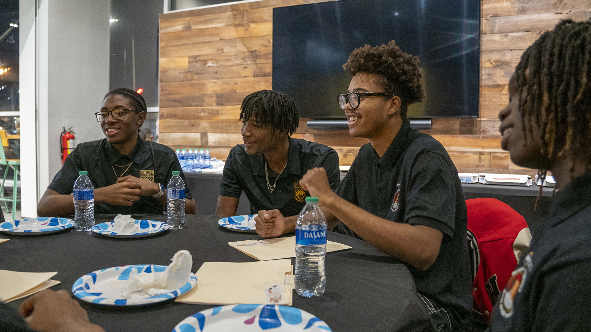 Four male students listen while sitting around one portion of a round table with water bottles and empty paper plates on its surface, in a room with a large monitor mounted on a wall with reclaimed wood paneling in front of plate-glass windows.