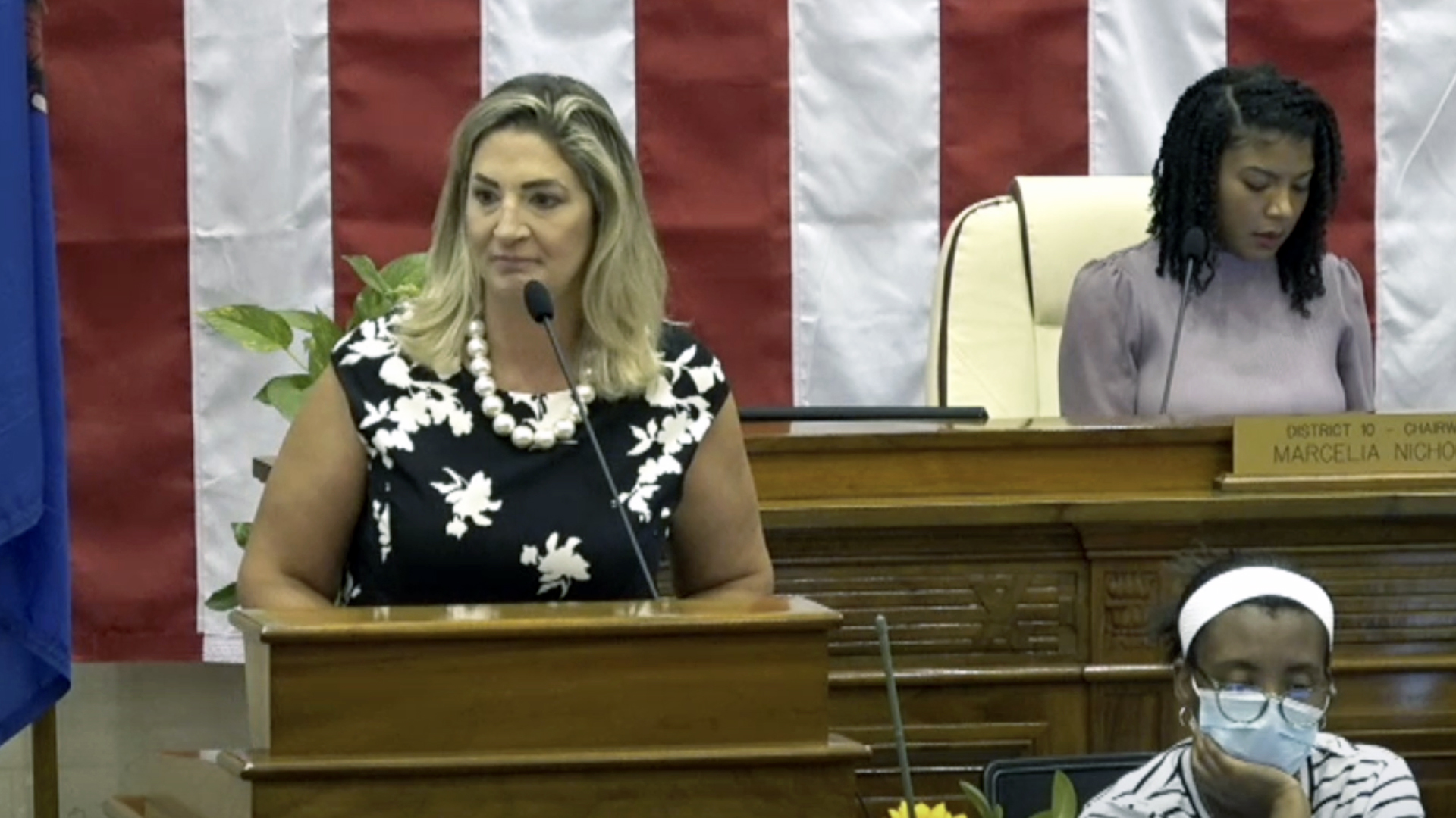 Margaret Daun speaks into a microphone while standing behind a wood podium, with two people to her side seated at a desk and legislative dais with wood carvings, with the vertical stripes of a large U.S. flag in the background.