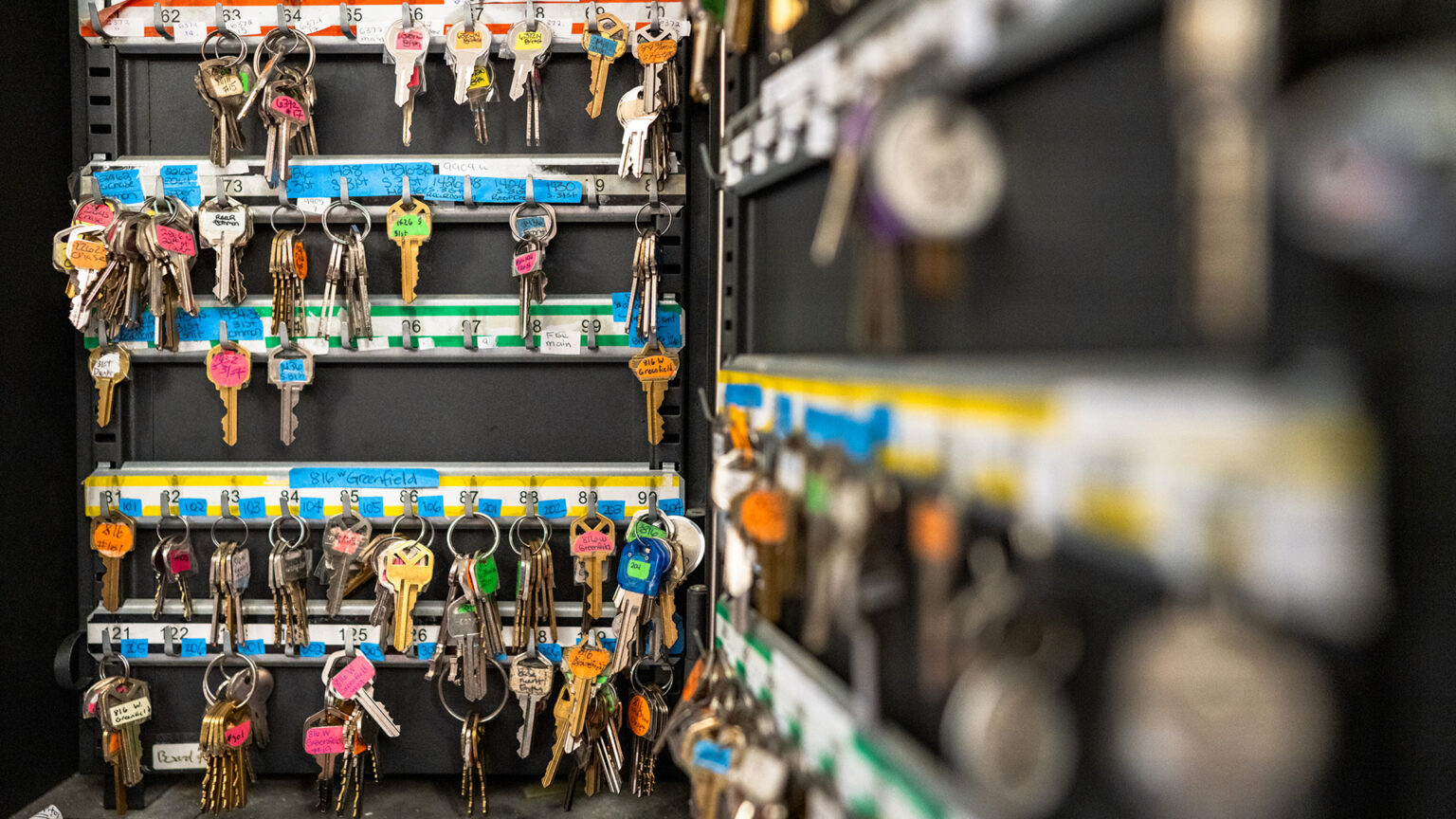 Multiple rings of keys with different color tape labels are arrayed in rows of hooks on two faces of a metal cabinet, with one in the background and another out-of-focus in the foreground.