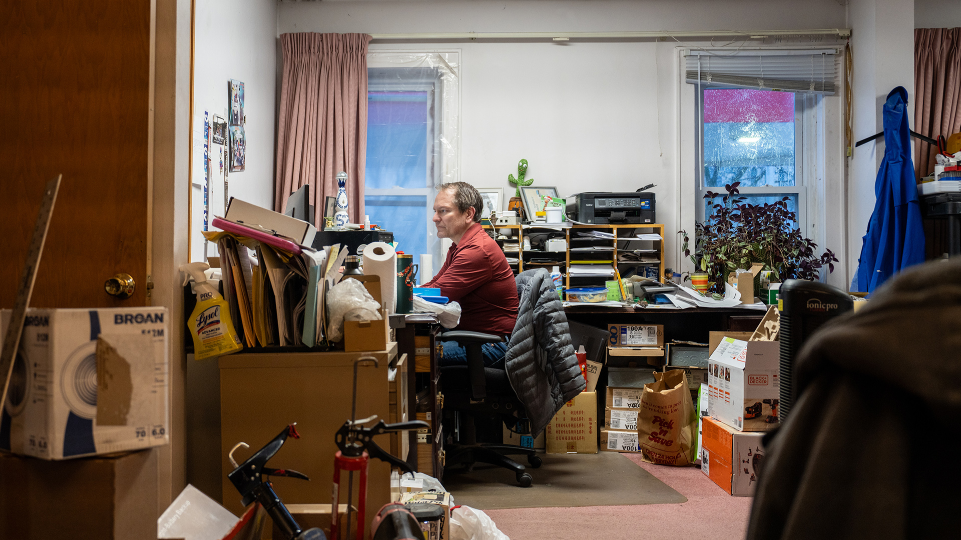 Sam Stair sits in a chair and faces a desk in a room with multiple stacks of boxes and other items and a table with a sorting shelf and other items on its surface against a wall with two windows with raised blinds.