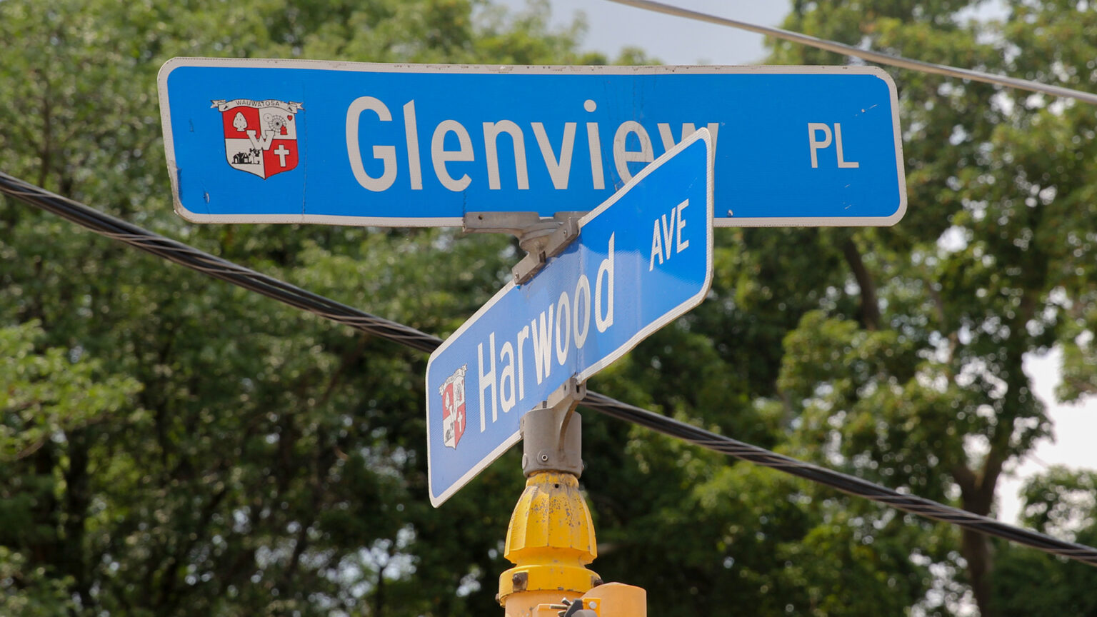 Two street signs with the Seal of Wauwatosa and the words Glenview PL and Harwood AVE stand at right angles to each other atop a metal pole, with power lines and out-of-focus trees in the background.