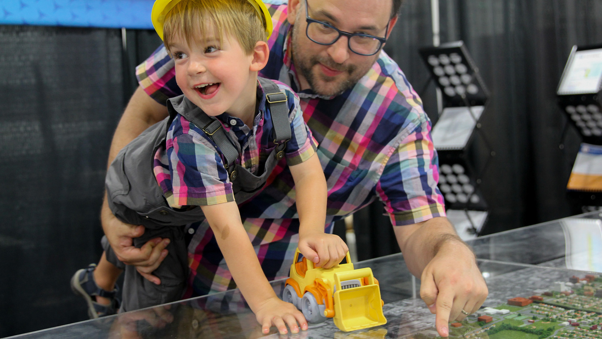 Robin Palm holds his son with his right arm while leaning over and pointing with his left hand toward a tabletop model display of a community under a transparent cover, with portable curtain room dividers in the background.