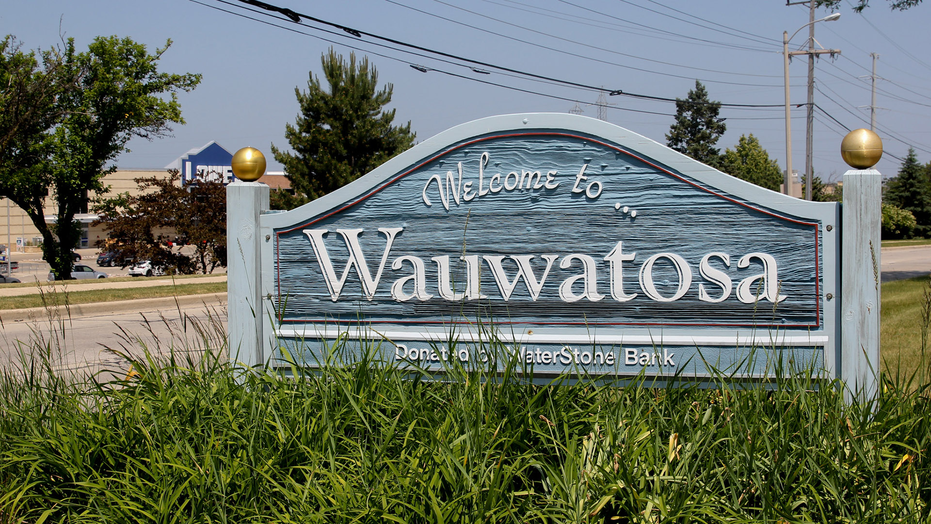 A carved wood sign in the form of a curved headboard with posts on either side features the words "Welcome to..." and "Wauwatosa," with plants at the bottom partially obscuring the words "Donated by WaterStone Bank," with a road, power lines, trees, buildings and a parking lot in the background.