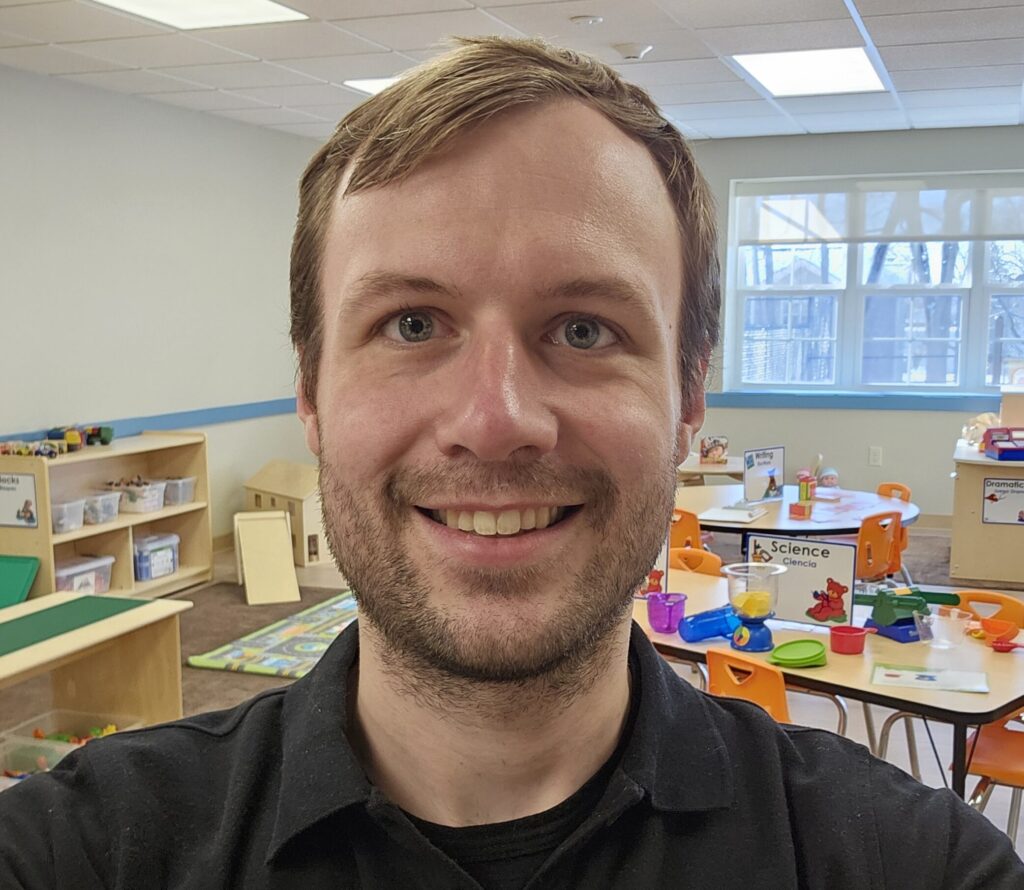 A teacher stands in an early learning classroom, smiling at the camera.