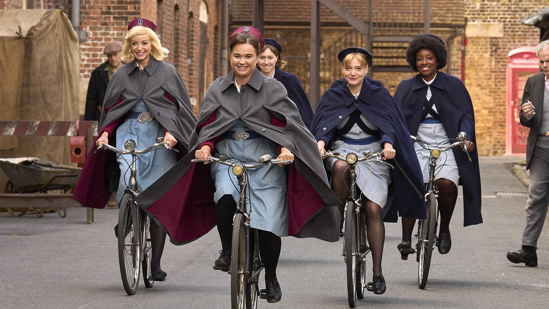 Five women dressed in blue nurse dresses, black tights, red caps and gray shawls ride bicycles in a group.