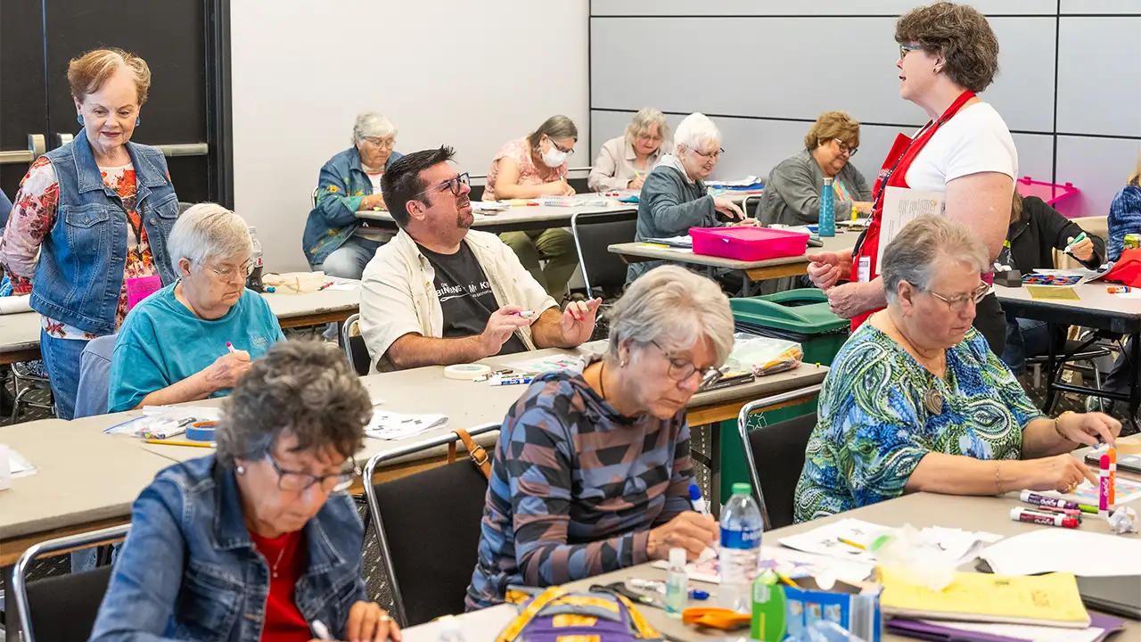 Older adults are engaged in a classroom setting, working on crafts and listening to an instructor at the front.