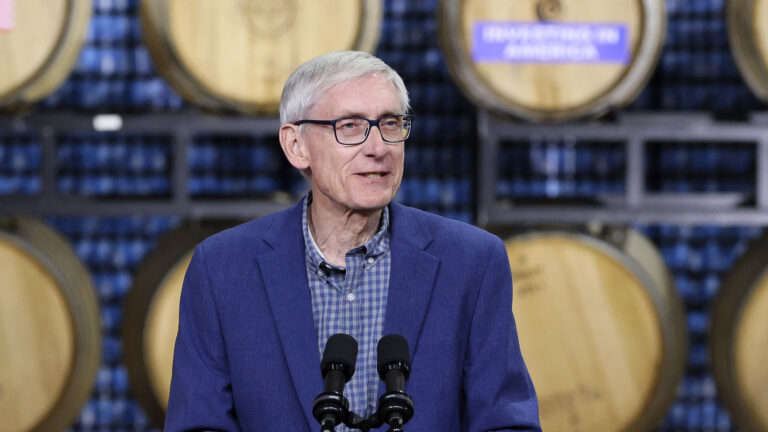 Tony Evers speaks into a pair of microphones while standing in front of shelves with wooden barrels stored on their sides.
