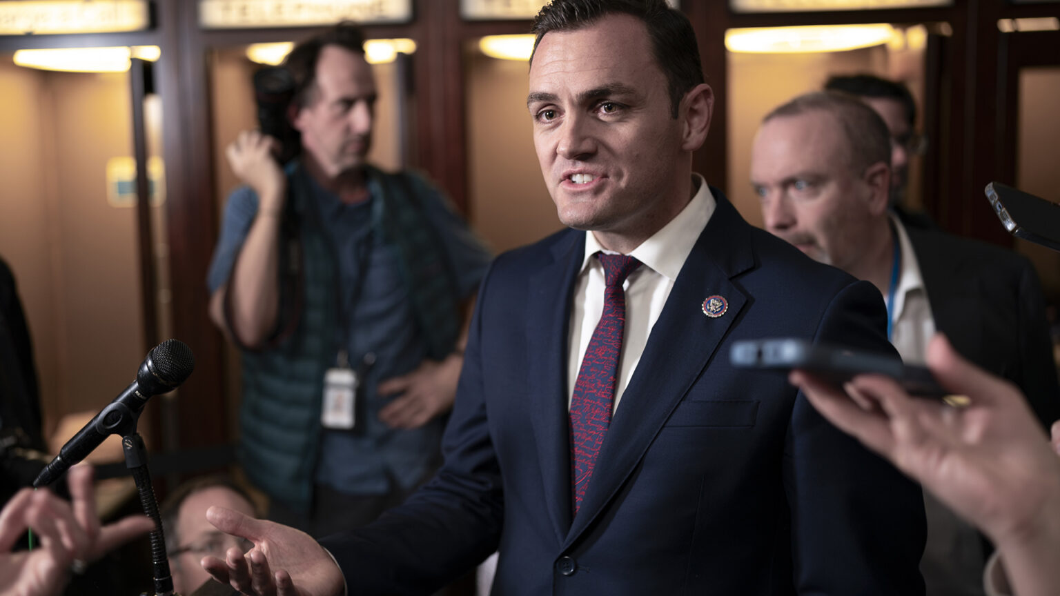 Mike Gallagher speaks while gesturing with his right hand, surrounded by reporters, including two holding cell phones to record the audio.