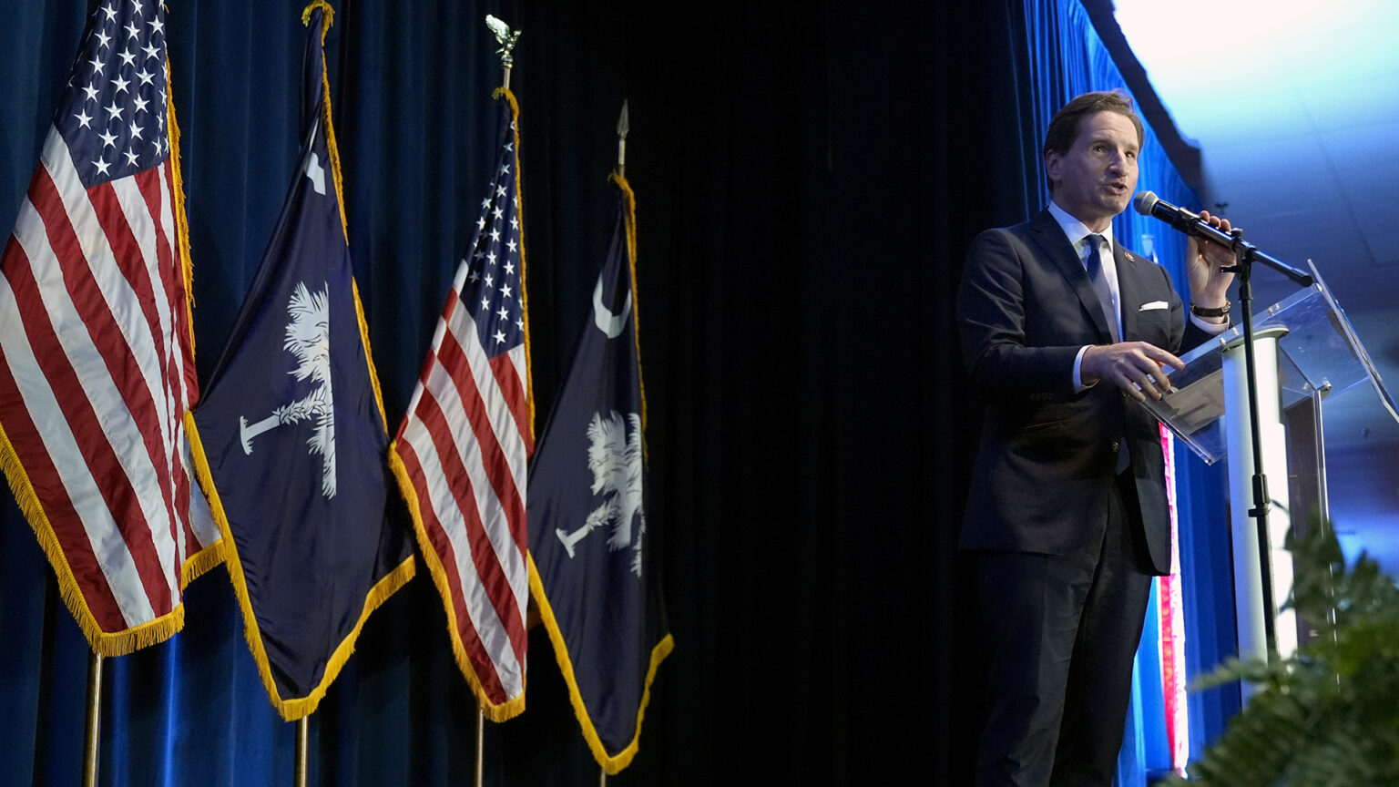 Dean Phillips speaks while holding a microphone mounted to a podium with a transparent surface, with an out-of-focus plant in the foreground and two South Carolina, two U.S. flags and a stage curtain in the background.