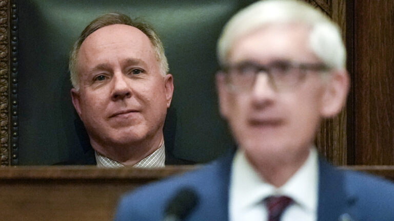 Robin Vos is seated in a high-backed chair behind Tony Evers, who is speaking (and out of focus) in the foreground.