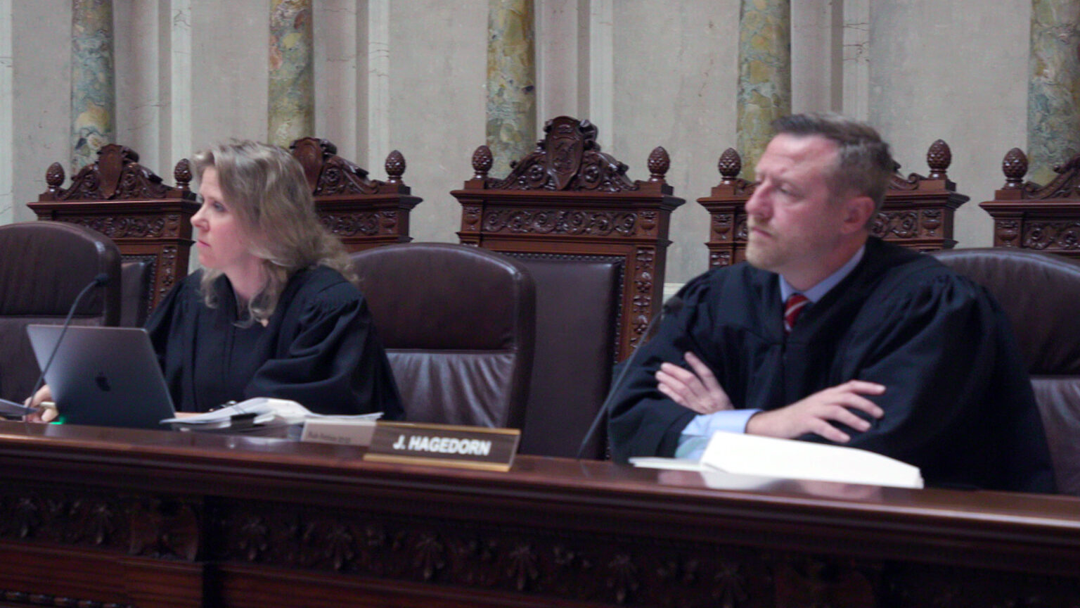 Rebecca Bradley and Brian Hagedorn sit in high-backed leather chairs at a judicial bench with a nameplate reading J. Hagedorn, with a row of empty high-backed wood and leather chairs behind them in a room with marble pillars and masonry.