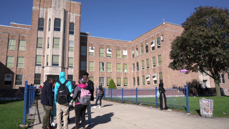 Students stand and walk along a concrete sidewalk with metal fences on either side in front of a multi-story brick building with multiple wings, with a tree on a lawn to the side.