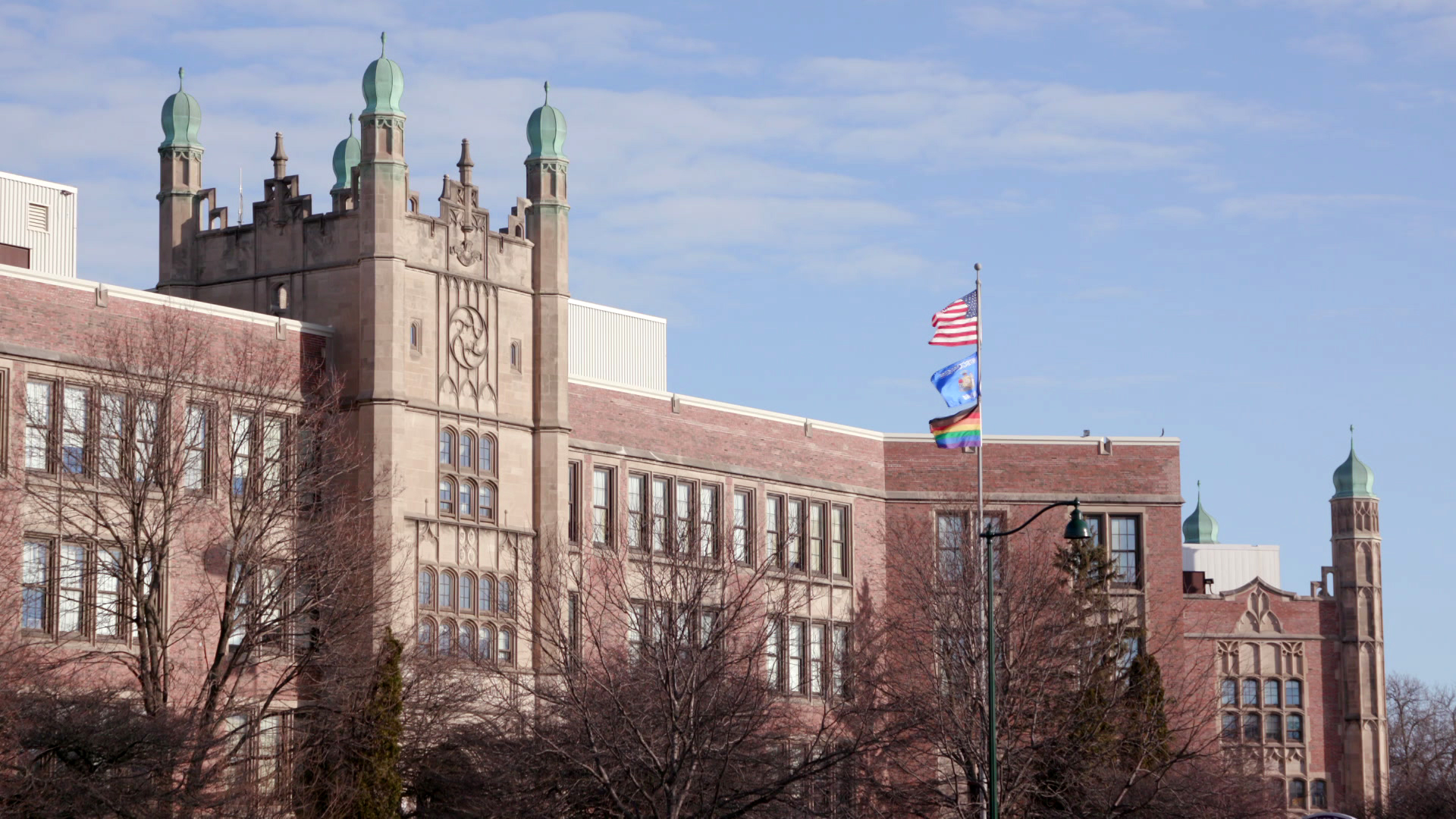 The U.S., Wisconsin and a Pride flag fly from a flagpole in front of a multi-story masonry and brick building with pillars on a central tower and on a wing topped by onion domes, along with HVAC machinery housing mounted on the roof, with leafless trees in the foreground.