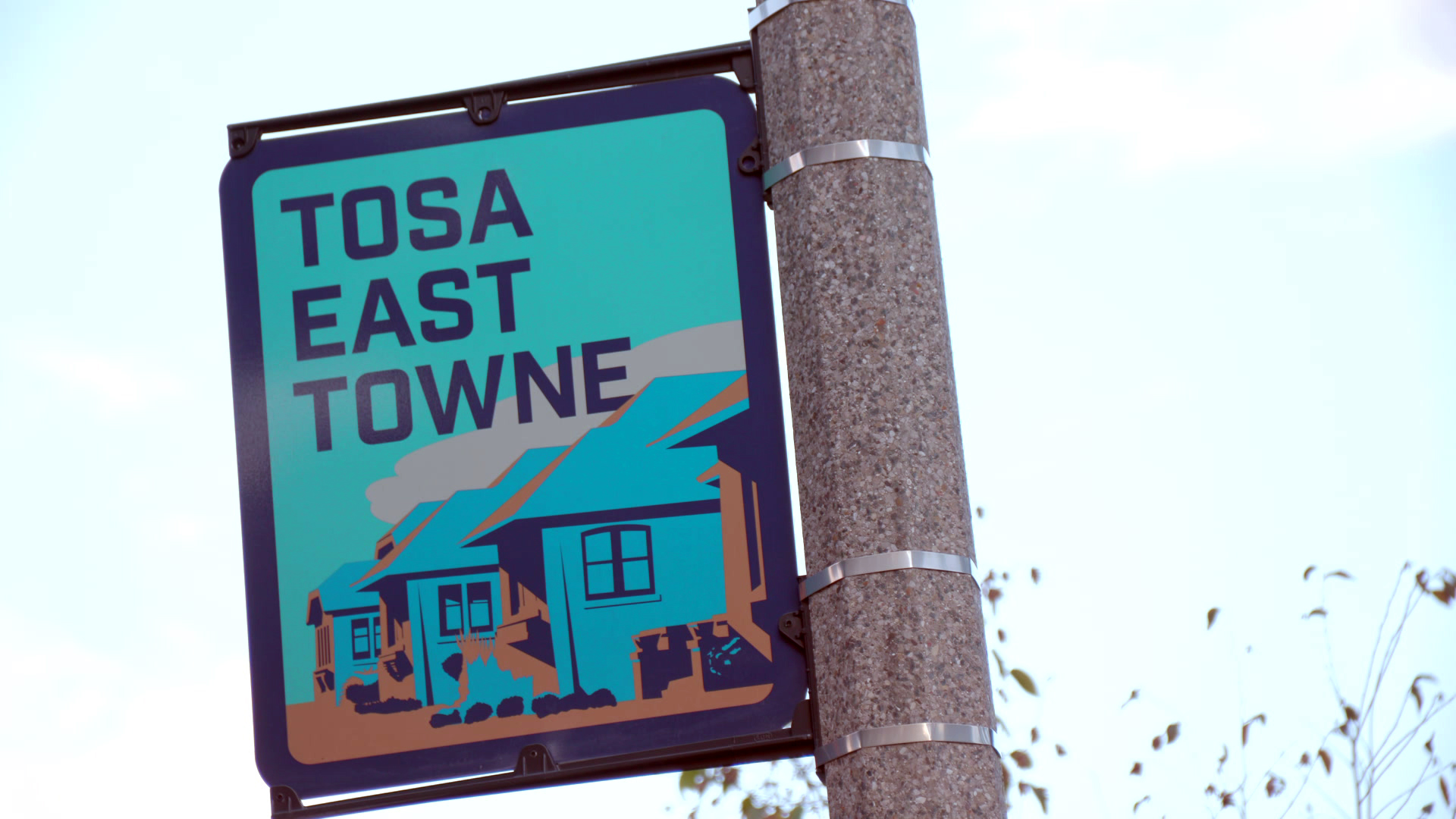 A sign with the words "Tosa East Towne" and a graphic of a row of houses is mounted with metal attachment rings to a stone pillar.
