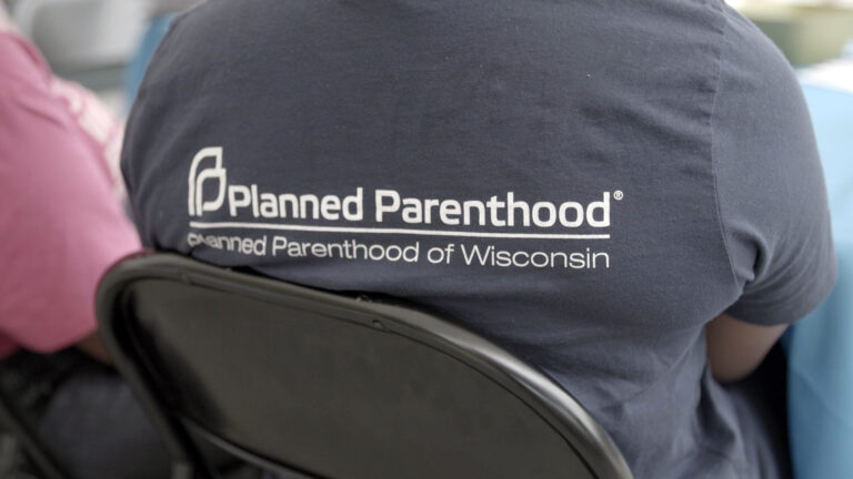 A person seated in a folding metal chair wears a t-shirt that has the Planned Parenthood wordmark and the words Planned Parenthood of Wisconsin on its back.