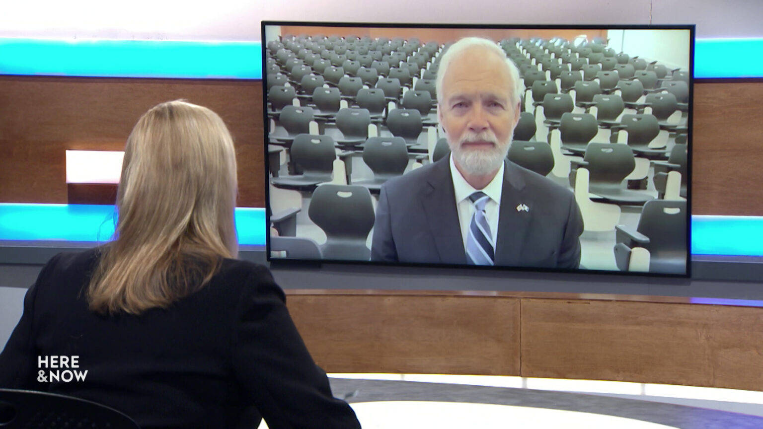 Frederica Freyberg sits at a desk on the Here & Now set and faces a video monitor showing an image of Ron Johnson.