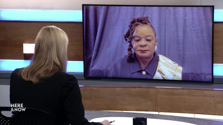 Frederica Freyberg sits at a desk on the Here & Now set and faces a video monitor showing an image of Gwen Moore.