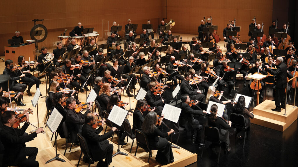 A shoulder view of the Madison Symphony Orchestra playing in Overture Hall as John DeMain conducts.