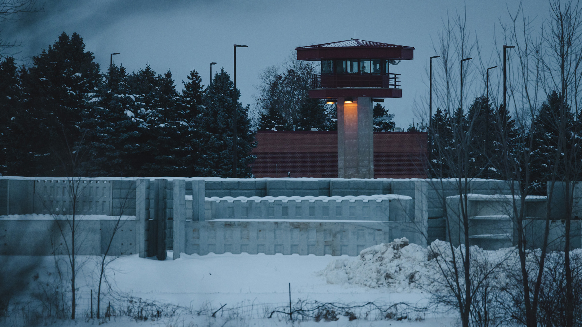 A prison guard tower build of concrete blocks and a metal observation post at top with windows on all sides stands above concrete walls and among light poles, with a snow covered field and leafless deciduous trees in the foreground, and snow-covered coniferous trees in the background.
