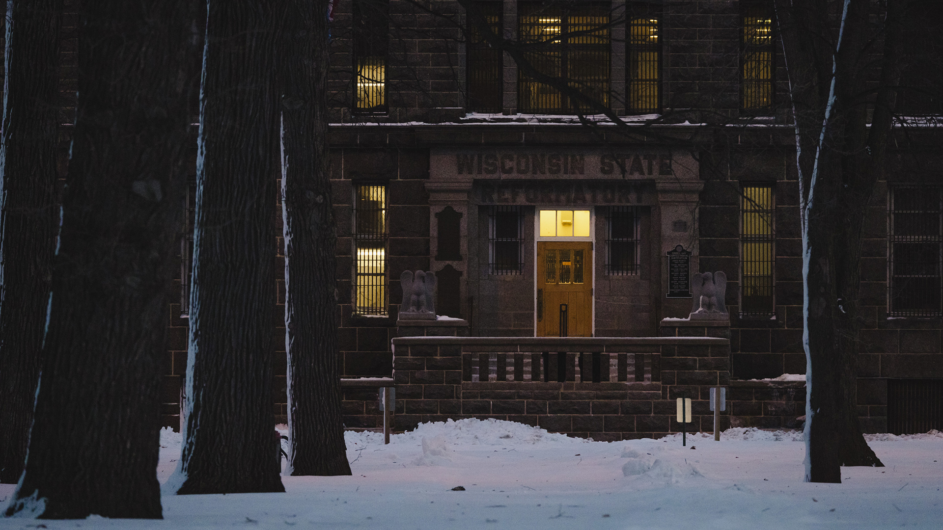 Light illuminates the interior to the entrance of a stone masonry building with a wood door and relief sign reading "Wisconsin State Reformatory," with light shining through bar-covered windows to either side and on a higher floor, with a snow-covered sidewalk and tree trunks in the foreground.