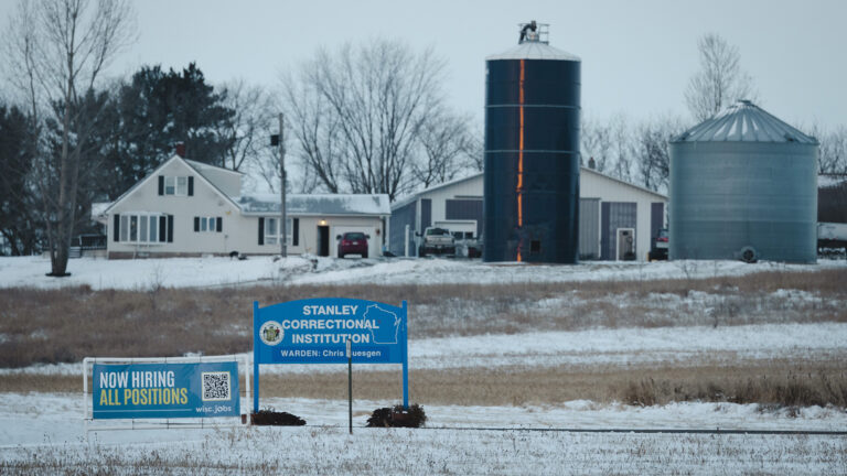 A temporary sign with the words Now Hiring All Positions and a QR code stands next to another sign showing the Great Seal of the State of Wisconsin, an outline of the state and the words Stanley Correctional Institution in a snow-covered agricultural field, with a house, two silos, other farm outbuildings and trees in the background.
