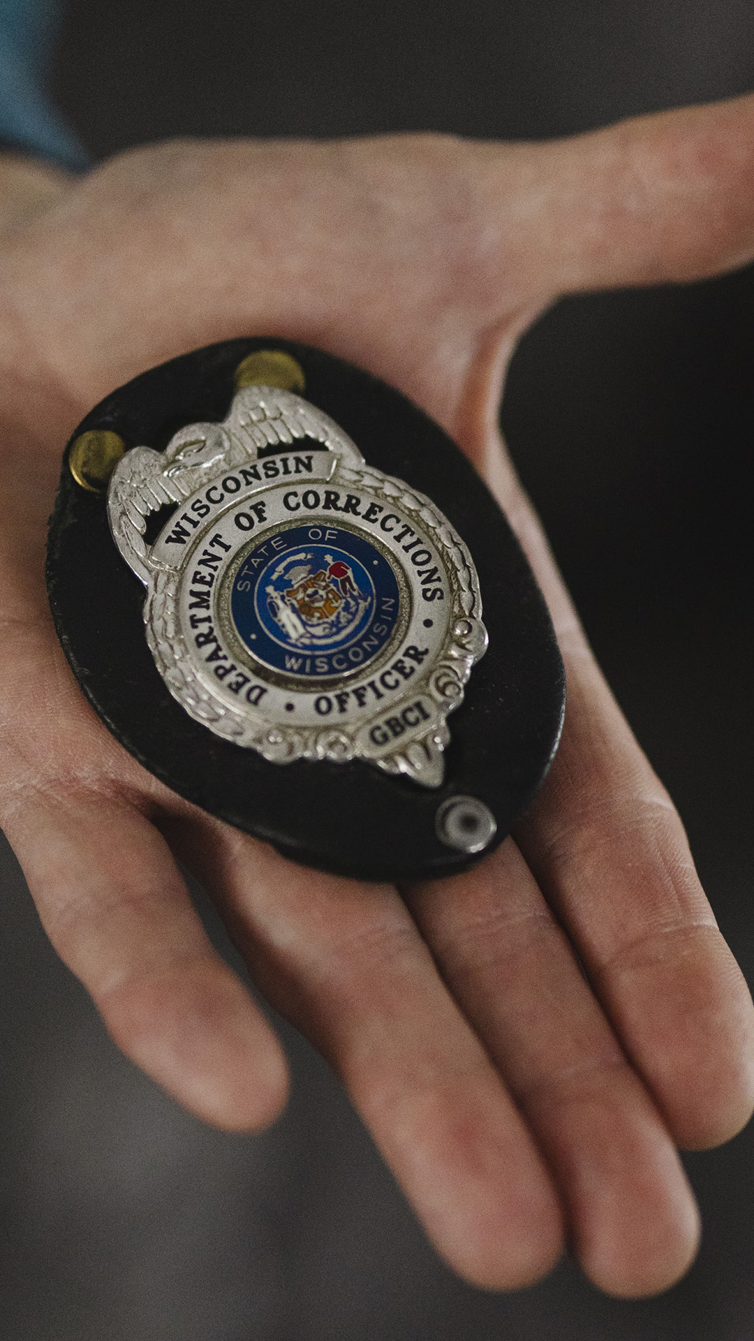 A left hand holds a law enforcement badge that reads "Wisconsin Department of Corrections" and "GBCI."