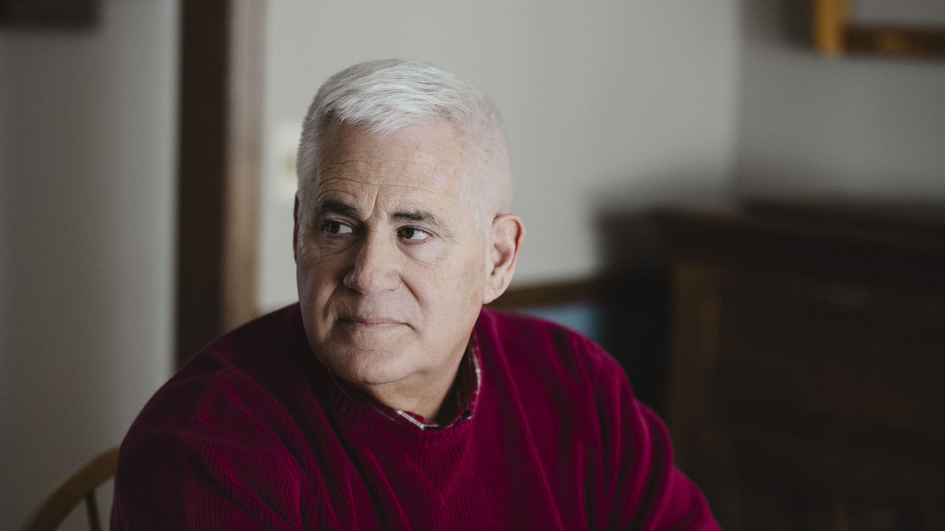 Ed Wall poses for a portrait while seated in a chair and looking to his right, in a room with out-of-focus wood furniture in the background.
