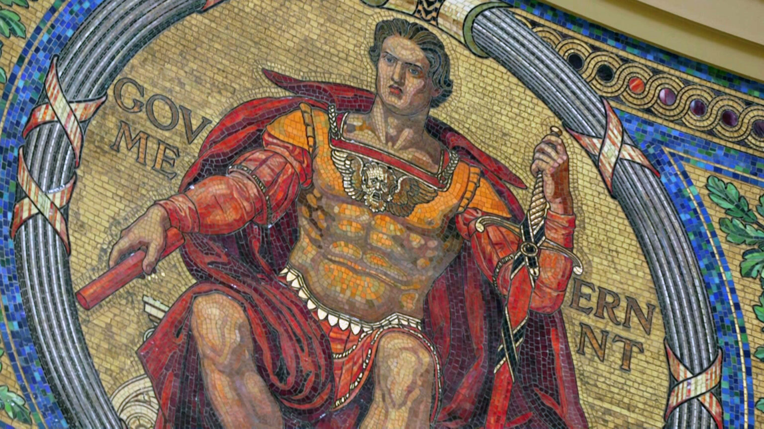 A mosaic depicting a Classical figure holding a sword and scroll and labeled Government is seen on a curved interior wall of a rotunda inside a building.