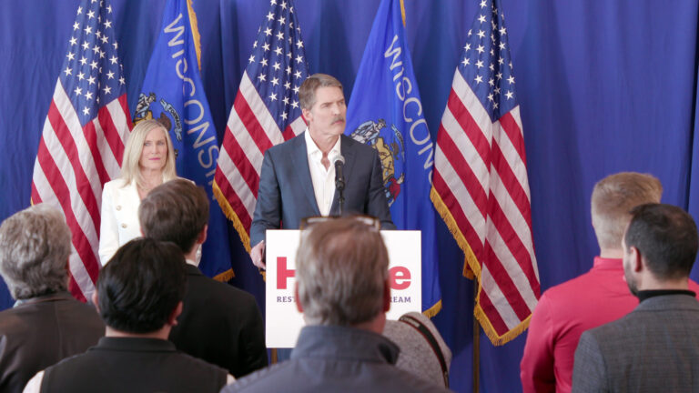 Eric Hovde speaks into a microphone mounted to a podium with another person standing to his side and others facing him in a room with a stage curtain and U.S. and Wisconsin flags in the background.
