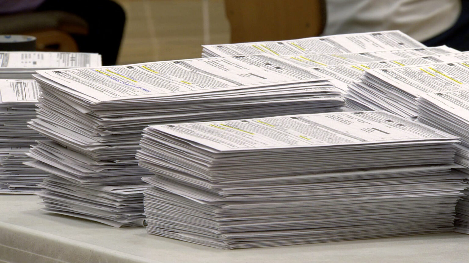 Multiple stacks of absente ballots envelopes with highlighted signature lines sit on a table.