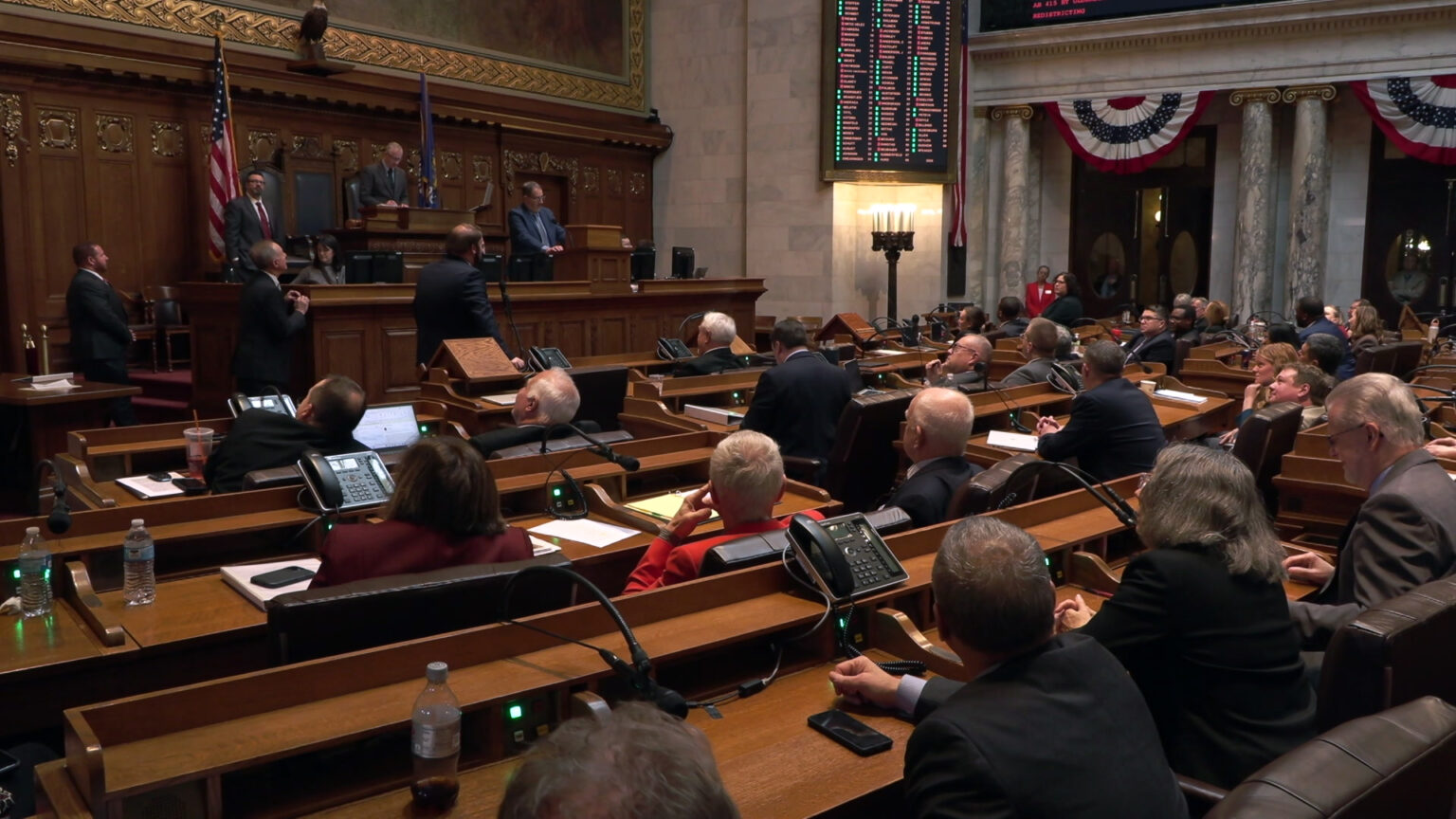 Legislators sit in high-backed leather chairs in multiple rows of desks facing a wood legislative dais where people are sitting and standing, with a U.S. and Wisconsin flags, a taxidermy bald eagle and large painting behind it, a digital vote register to the side of the dais, in a room with marble pillars and masonry.