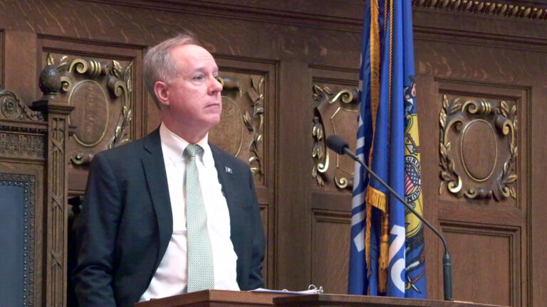 Robin Vos stands and faces a microphone, with a high-backed leather and wood chair and a Wisconsin flag behind him, in a room with wood paneling featuring carved relief crests.
