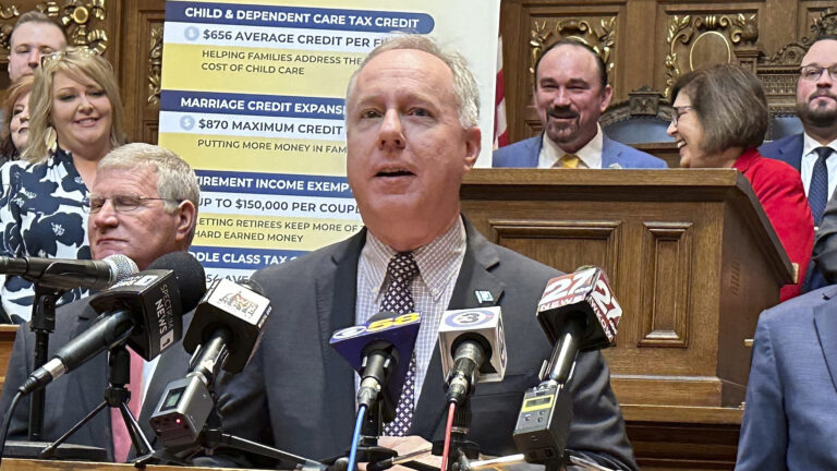 Robin Vos speaks into multiple microphones with the flags of different media organizations as other people stand on either side and on a higher tier of a legislative dais, with a sign with multiple bullet points in the background of a room with carved wood wall panels.