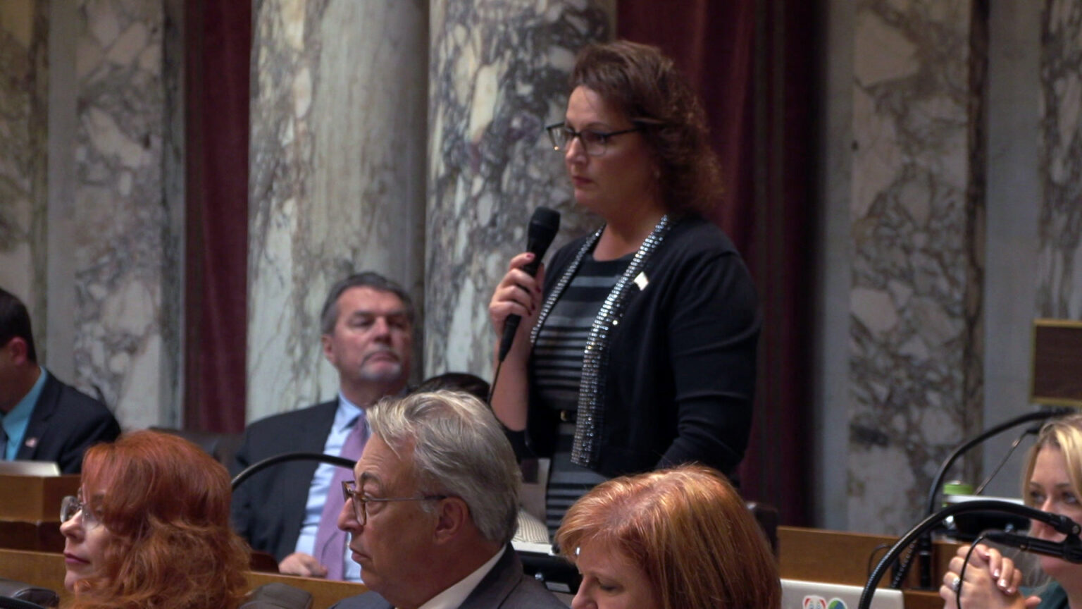 Amy Binsfield speaks into a microphone she is holding in her right hand while standing among other people seated in rows of wood desks, in a room with marble masonry.