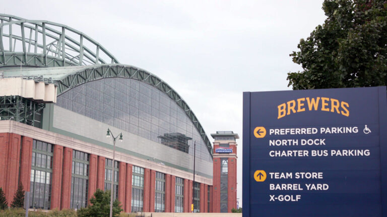 A sign labeled Brewers and with arrows pointing toward different locations stands under a tree and in front of a baseball stadium with a closed retractable roof under a cloudy sky.