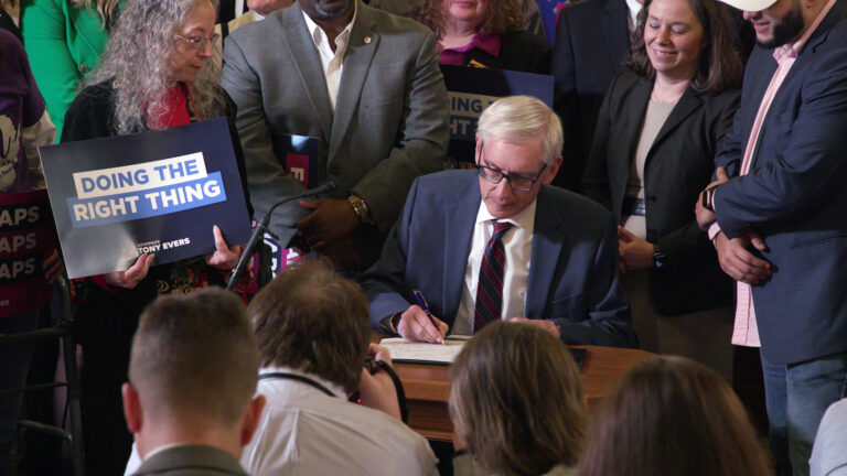 Tony Evers signs a document with a pen in his right hand while seated at a wooden desk, with other people standing around and behind him, with several holding printed signs reading Doing The Right Thing, in a room with other seated people facing him.