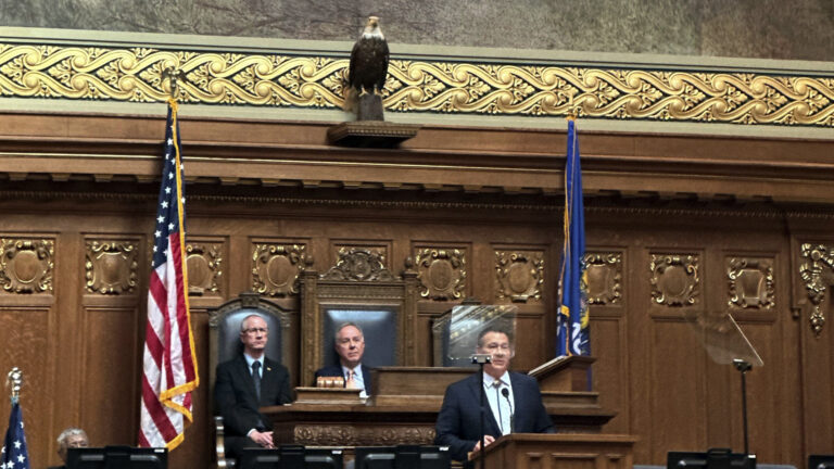 James Crawford speaks into a microphone while standing behind a wood podium and facing two teleprompter mirrors, with Robin Vos and others seated behind him in high-backed leather and wood chairs on a legislative dais, with a taxidermy bald eagle on display above a wall with wood paneling and carved crests and below a large painting with a gilt frame.