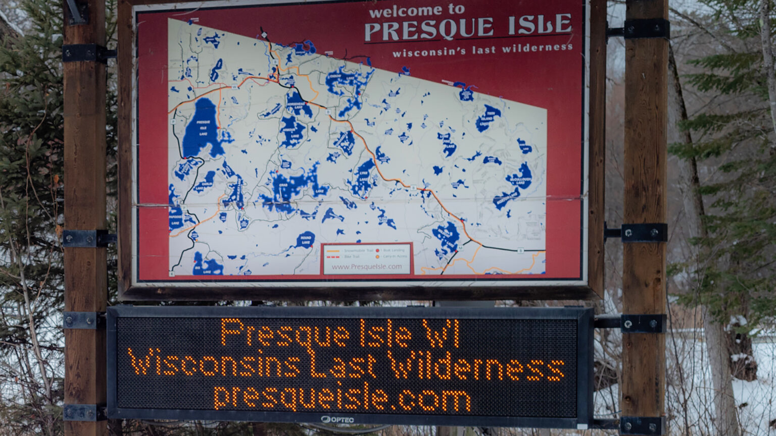 A sign with the words welcome to Presque Isle and wisconsin's last wilderness shows the outlines of a municipality with markers for roads and labels for bodies of water is placed between two wood posts, with an electronic sign below showing the words Presque Isle WI, Wisconsins Last Wilderness and presqueilse.com, with trees and snowbanks in the background.