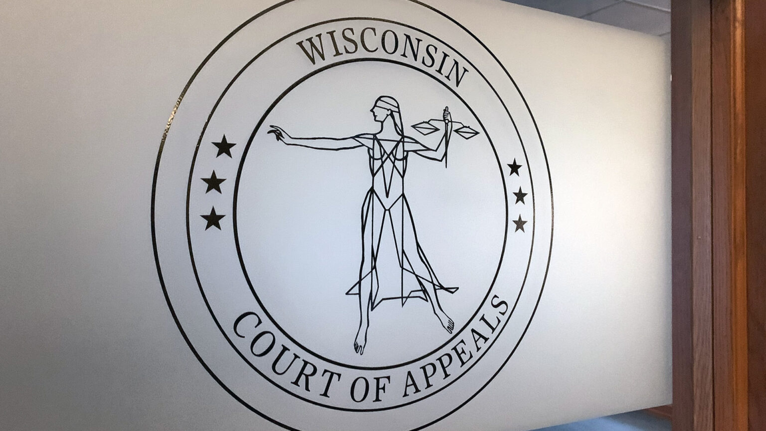 A graphic of the seal of the Wisconsin Court of Appeals, which consists of a line drawing of Lady Justice with three stars on either side and the words Wisconsin and Court of Appeals above and below, respectively, within multiple concentric circles, is visible on a frosted portion of large interior window encased within a wood window frame.