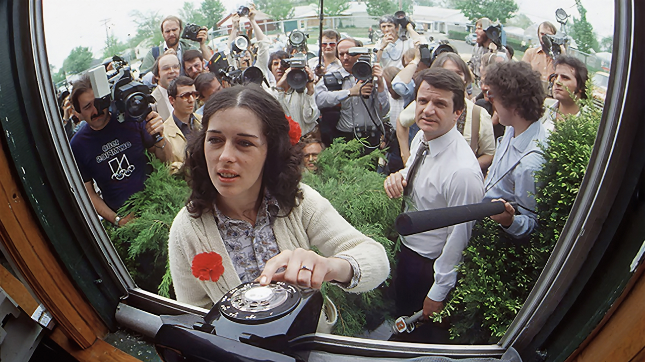 A crowd of cameramen surround a woman as she taps a rotary dial phone at an office window.