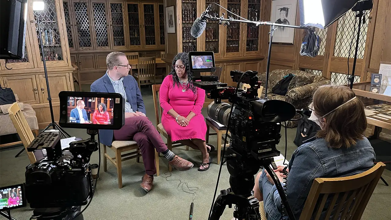Two people in an interview setup with video recording equipment in a room with wood-paneled walls and bookshelves.