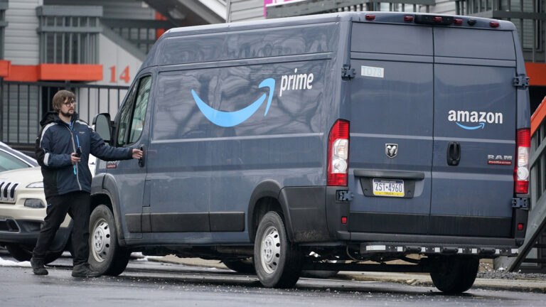 A delivery driver holds the driver's side door handle of a high roof cargo van with the Amazon arrow logo and the words prime and amazon, parked along the side of a road with other parked vehicles and buildings in the background.